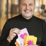 Holger Bramsiepe lachend mit bunten Postkarten in der Hand und schwarzem Rolli an