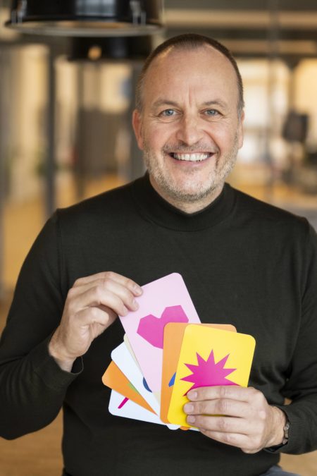 Holger Bramsiepe lachend mit bunten Postkarten in der Hand und schwarzem Rolli an